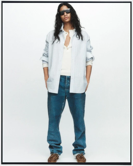 Man in unbuttoned blue striped shirt, white t-shirt, blue jeans, and brown shoes stands in a full shot against a white backdrop.