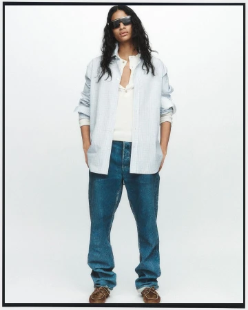 Man in unbuttoned blue striped shirt, white t-shirt, blue jeans, and brown shoes stands in a full shot against a white backdrop.