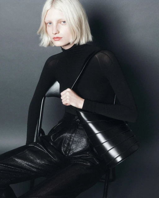 Blonde-haired woman in black turtleneck seated and holding a glossy black purse against a dark gray studio backdrop in a fashion editorial shot.