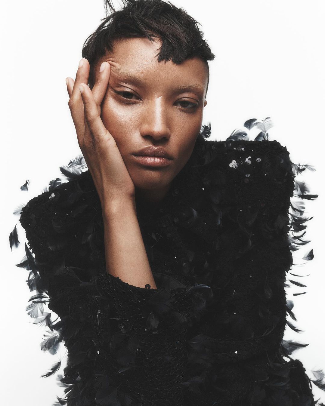 Close-up studio portrait of a young adult female model with short dark hair and dark eyes, wearing a black feathered top against white.