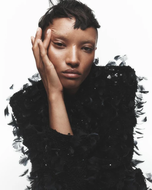 Close-up studio portrait of a young adult female model with short dark hair and dark eyes, wearing a black feathered top against white.