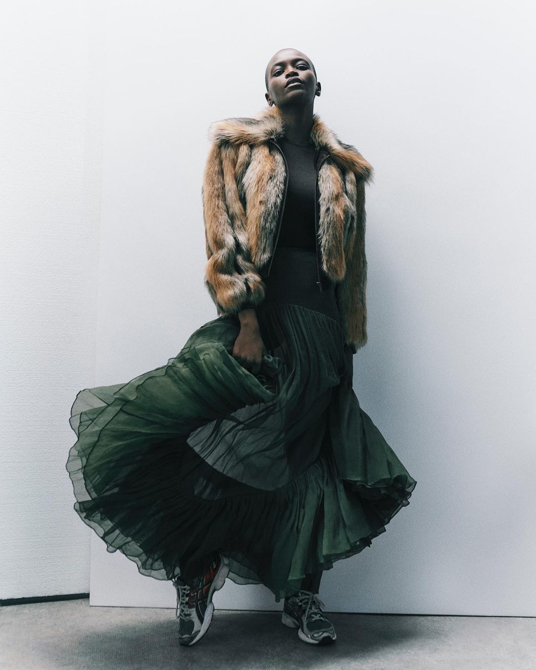 Woman in fur jacket and green skirt poses in a fashion shot against a white background, showcasing stylish attire.