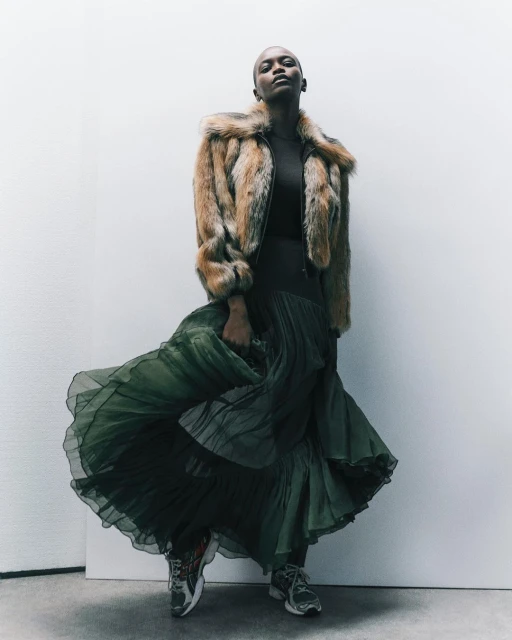 Woman in fur jacket and green skirt poses in a fashion shot against a white background, showcasing stylish attire.