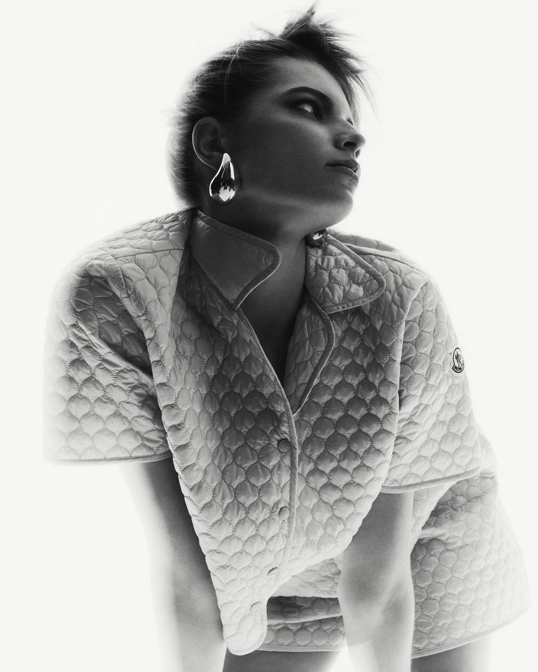 Woman with short hair wearing an open quilted jacket and silver teardrop earrings poses against a bright white background in studio shot.