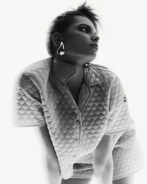 Woman with short hair wearing an open quilted jacket and silver teardrop earrings poses against a bright white background in studio shot.