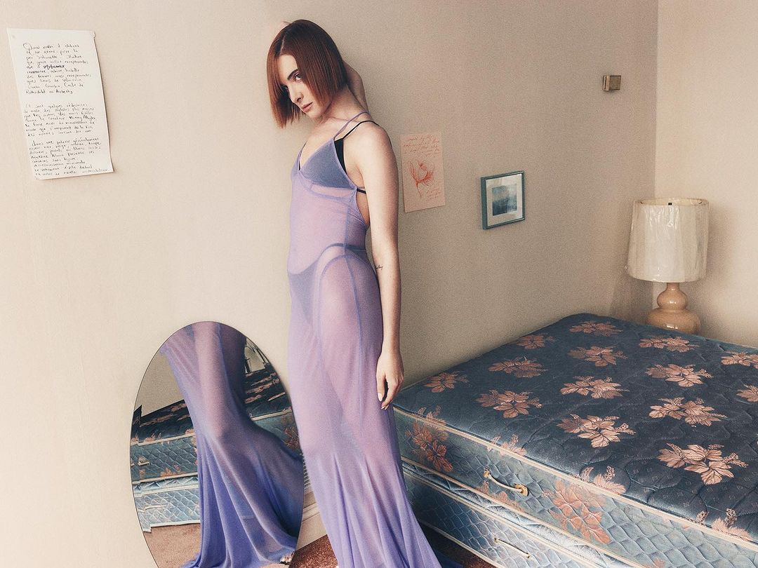 Adult female poses in a sheer lilac dress in a room with a bed, mirror, framed art, and a handwritten page on the wall. 