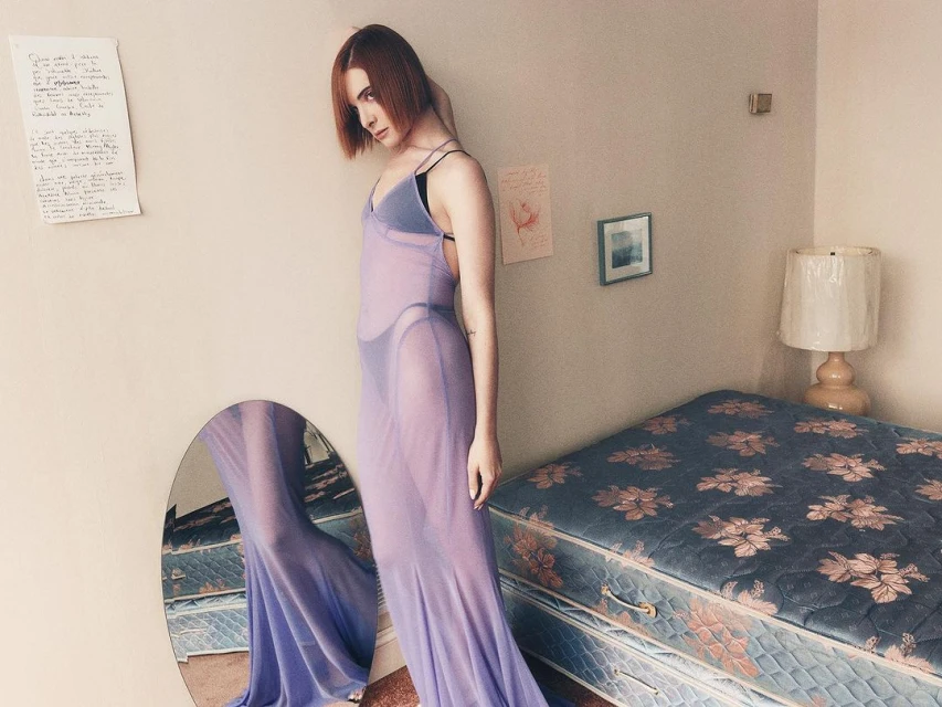 Adult female poses in a sheer lilac dress in a room with a bed, mirror, framed art, and a handwritten page on the wall. 
