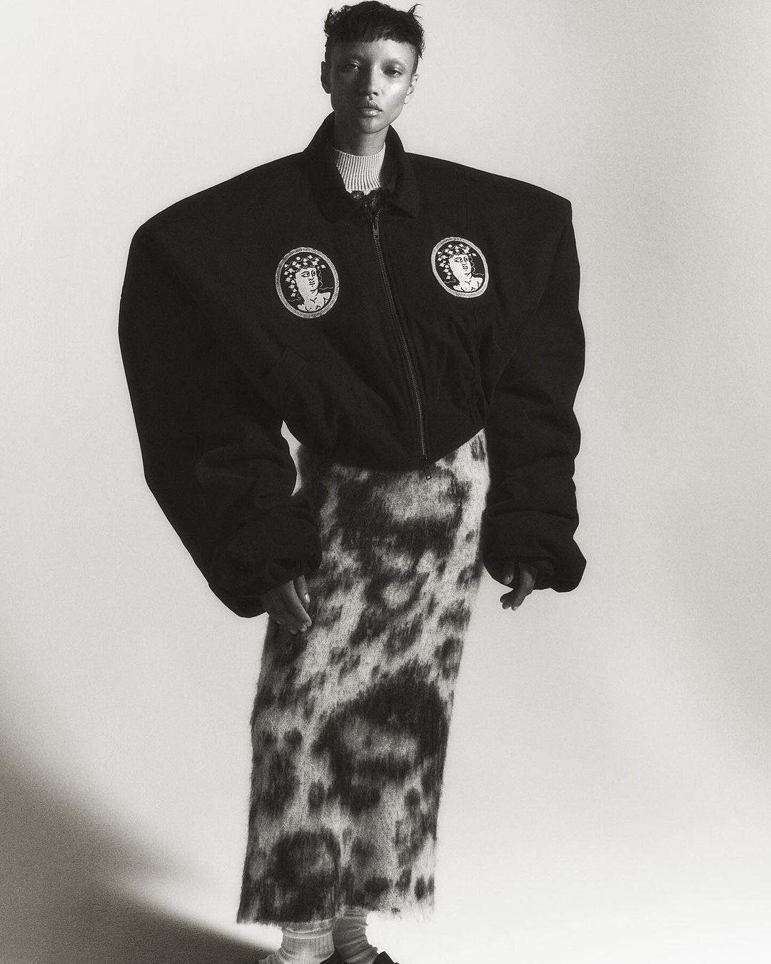 Monochrome fashion shot of a slender person with a buzzcut in an oversized black jacket and patterned skirt posing in a white studio.