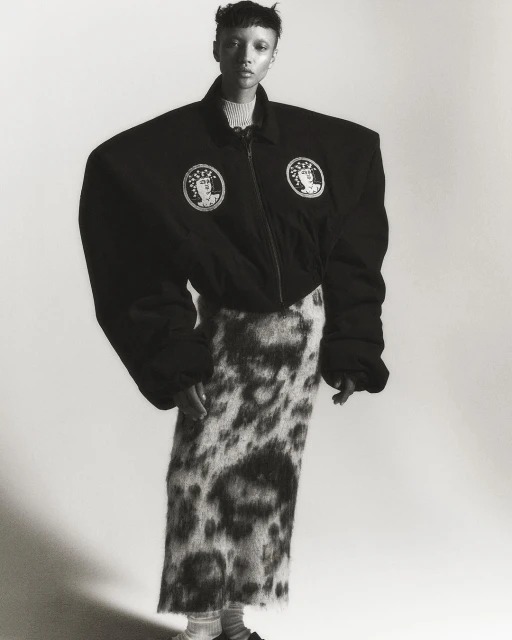Monochrome fashion shot of a slender person with a buzzcut in an oversized black jacket and patterned skirt posing in a white studio.