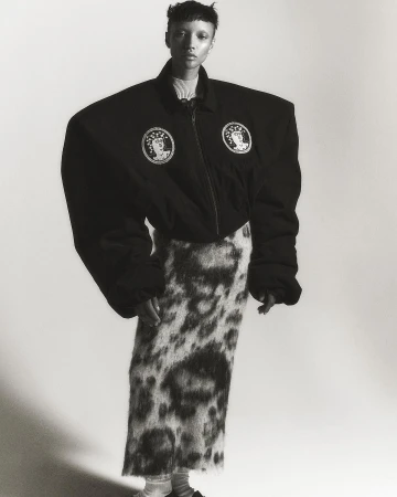 Monochrome fashion shot of a slender person with a buzzcut in an oversized black jacket and patterned skirt posing in a white studio.