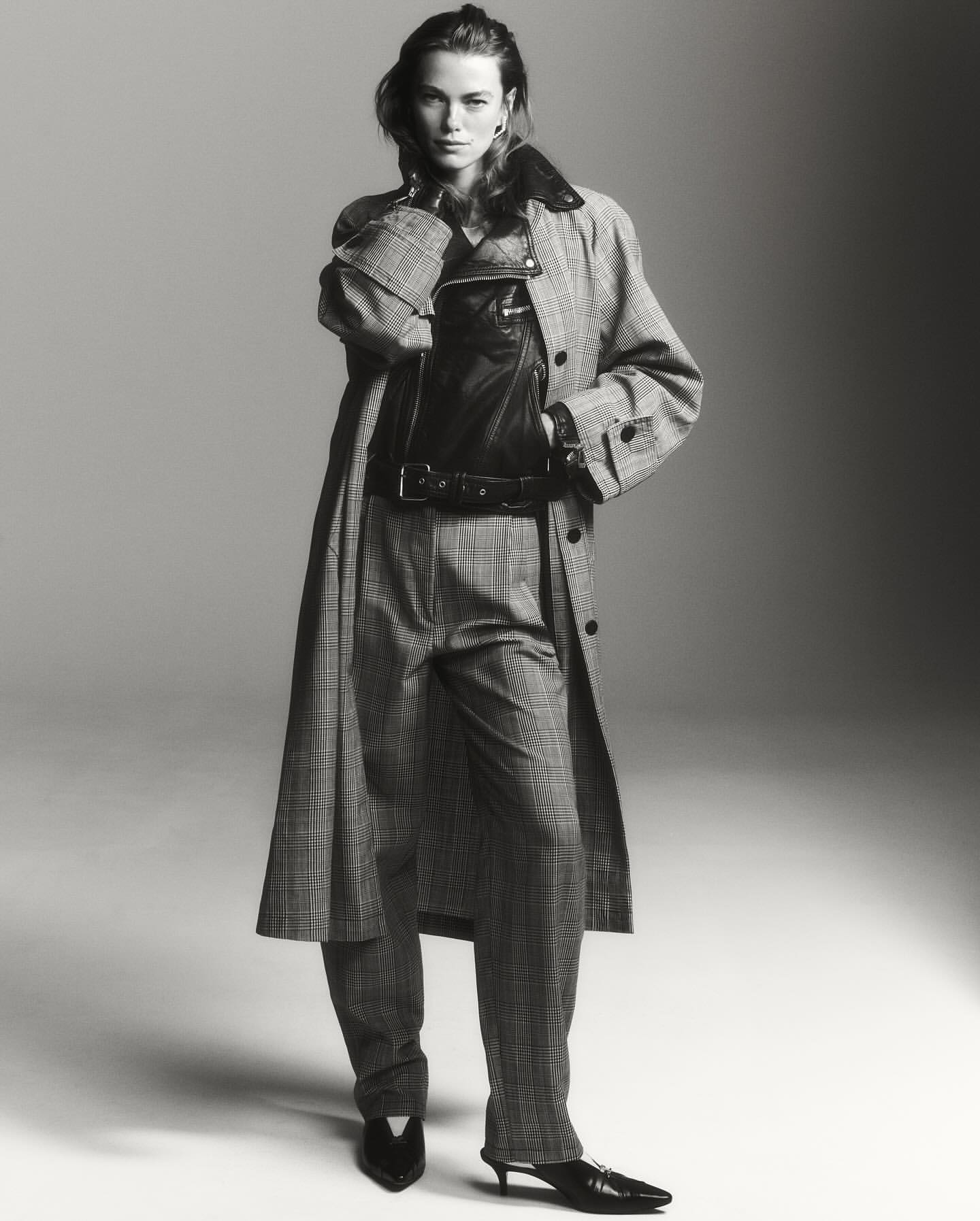 A woman in a patterned coat and pants poses in a studio. She is also wearing a leather vest. The image is monochrome, and the lighting is soft.