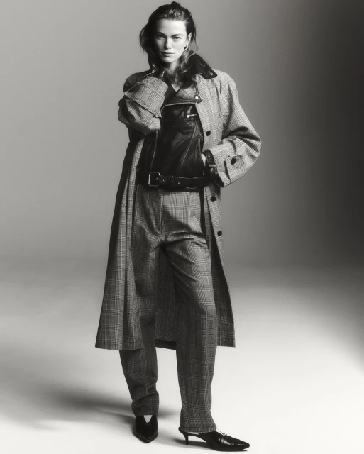 A woman in a patterned coat and pants poses in a studio. She is also wearing a leather vest. The image is monochrome, and the lighting is soft.