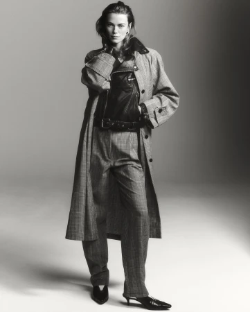 A woman in a patterned coat and pants poses in a studio. She is also wearing a leather vest. The image is monochrome, and the lighting is soft.