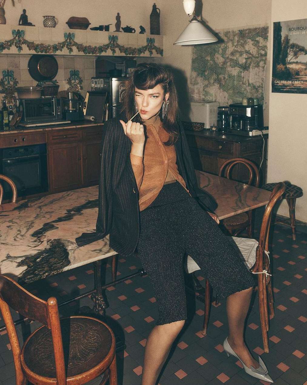 Brunette woman in a pinstripe blazer and culottes sits at a table in a kitchen, holding a toothpick and winking at the camera.