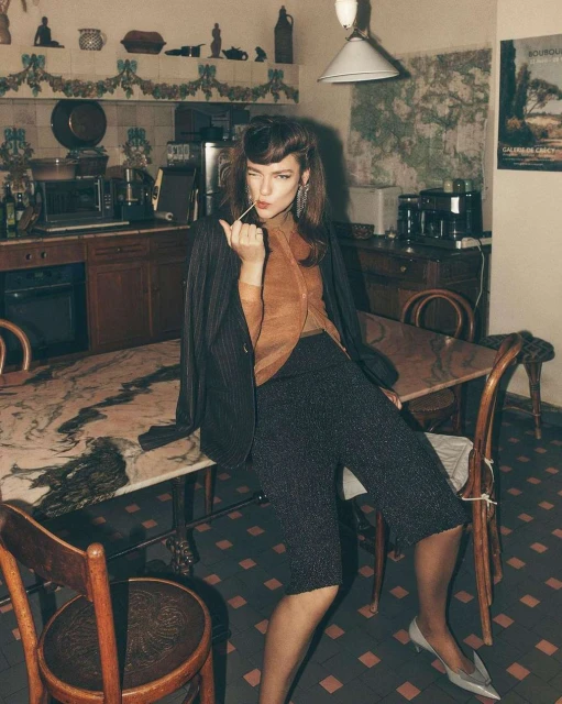 Brunette woman in a pinstripe blazer and culottes sits at a table in a kitchen, holding a toothpick and winking at the camera.