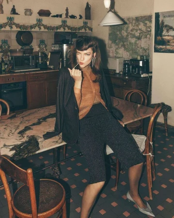 Brunette woman in a pinstripe blazer and culottes sits at a table in a kitchen, holding a toothpick and winking at the camera.