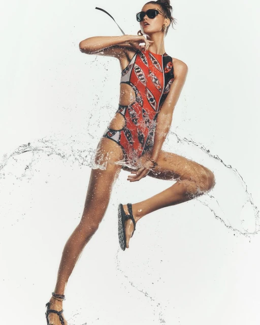 Athletic woman in a patterned swimsuit jumps with water splashing around her against a bright white background wearing sunglasses and sandals.