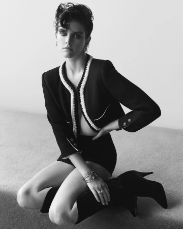 A fashionable woman with short hair poses kneeling indoors. She is wearing a black jacket, shorts and knee high boots against a neutral background.