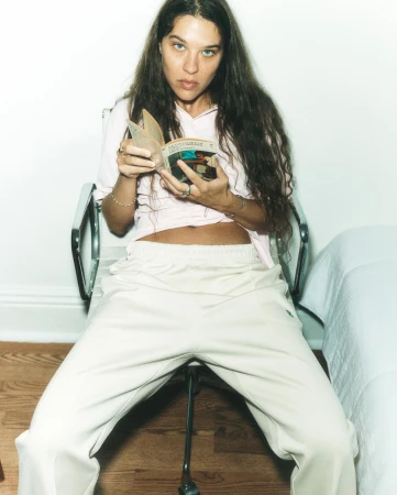 Woman with long dark curly hair seated in a chair reading a small book wearing a light pink top and off-white trousers.