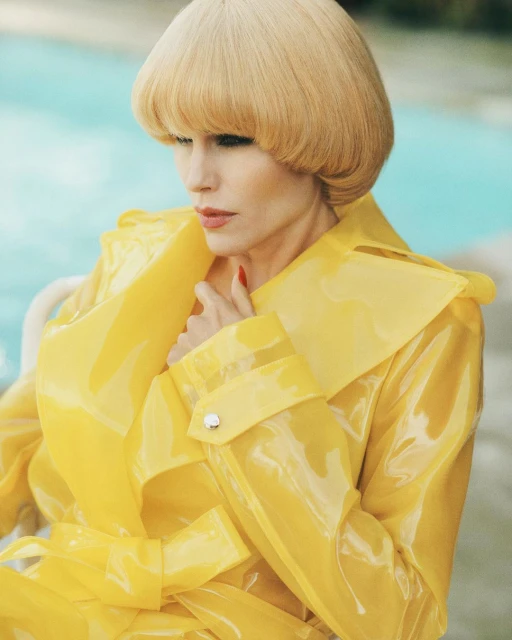 Woman in a glossy yellow trench coat poses poolside, looking downward, with a modern blonde bowl cut and red nails.