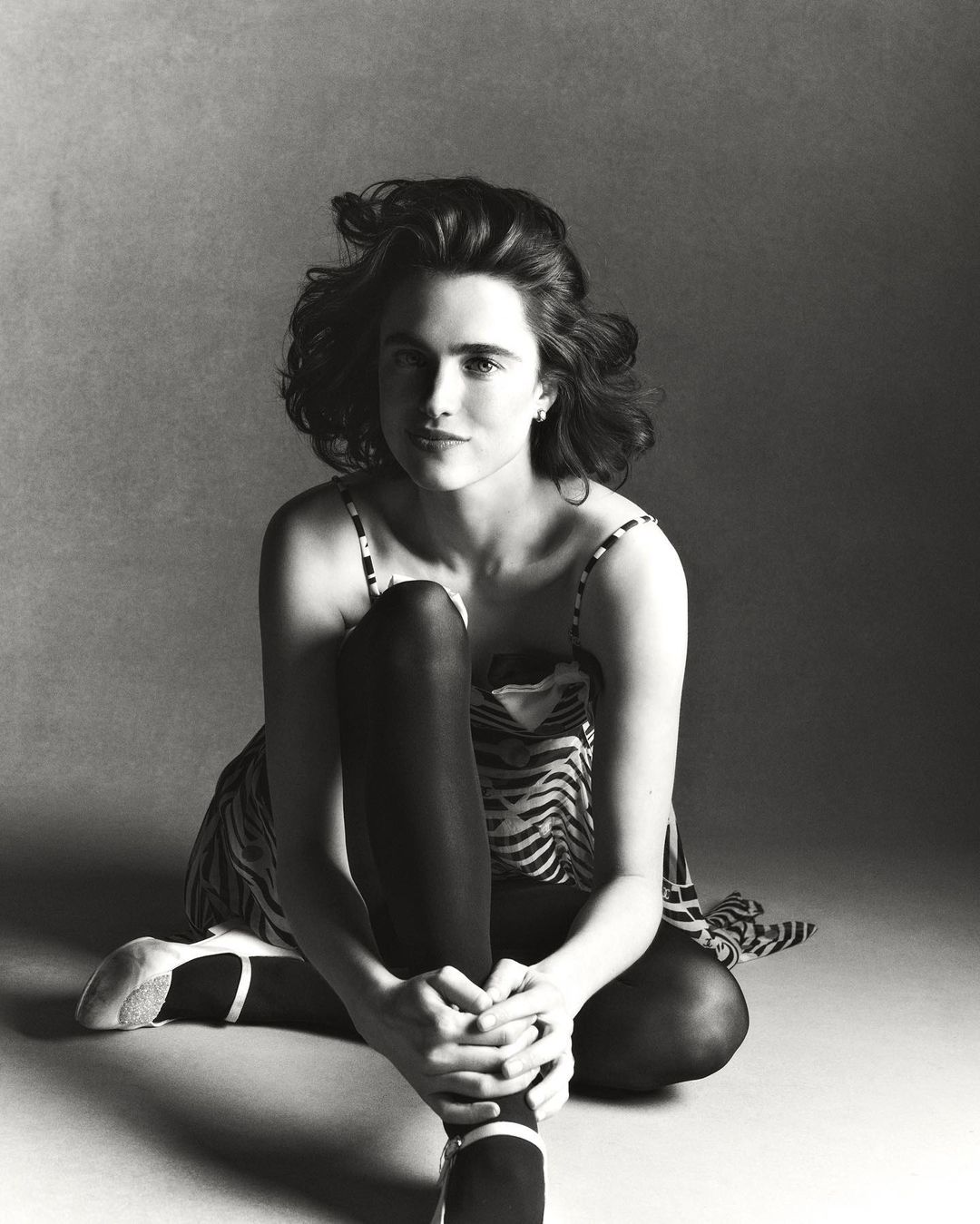 Monochrome studio portrait of a woman with curly hair sitting cross-legged wearing a patterned dress and ballet flats against a grey backdrop.