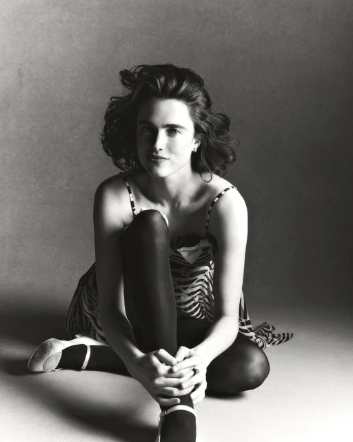 Monochrome studio portrait of a woman with curly hair sitting cross-legged wearing a patterned dress and ballet flats against a grey backdrop.