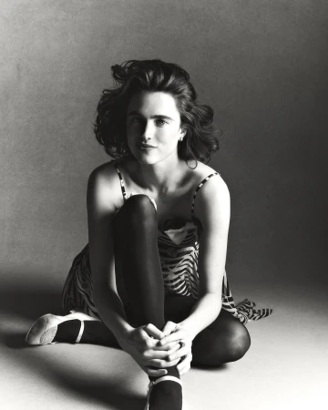 Monochrome studio portrait of a woman with curly hair sitting cross-legged wearing a patterned dress and ballet flats against a grey backdrop.
