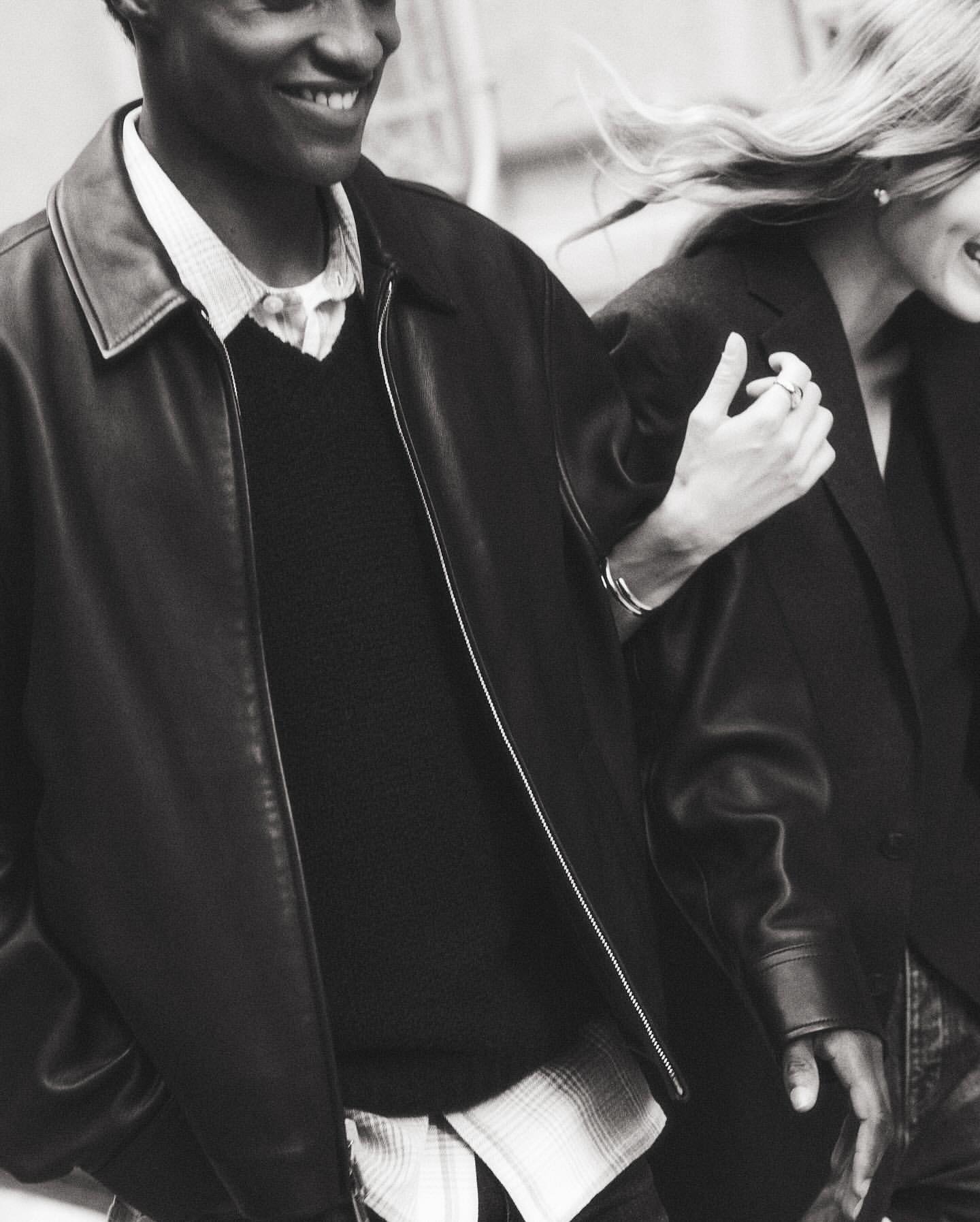 Monochrome shot of a smiling Black man in a leather jacket, arm-in-arm with a smiling white woman, both wearing dark jackets.