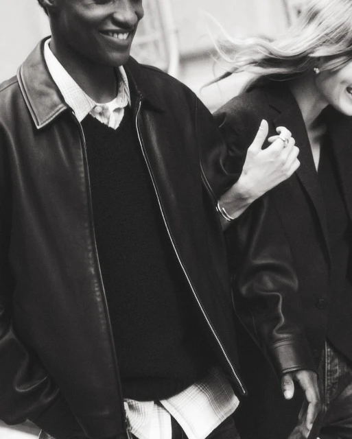 Monochrome shot of a smiling Black man in a leather jacket, arm-in-arm with a smiling white woman, both wearing dark jackets.