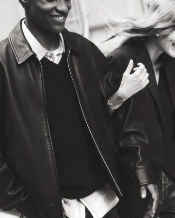 Monochrome shot of a smiling Black man in a leather jacket, arm-in-arm with a smiling white woman, both wearing dark jackets.