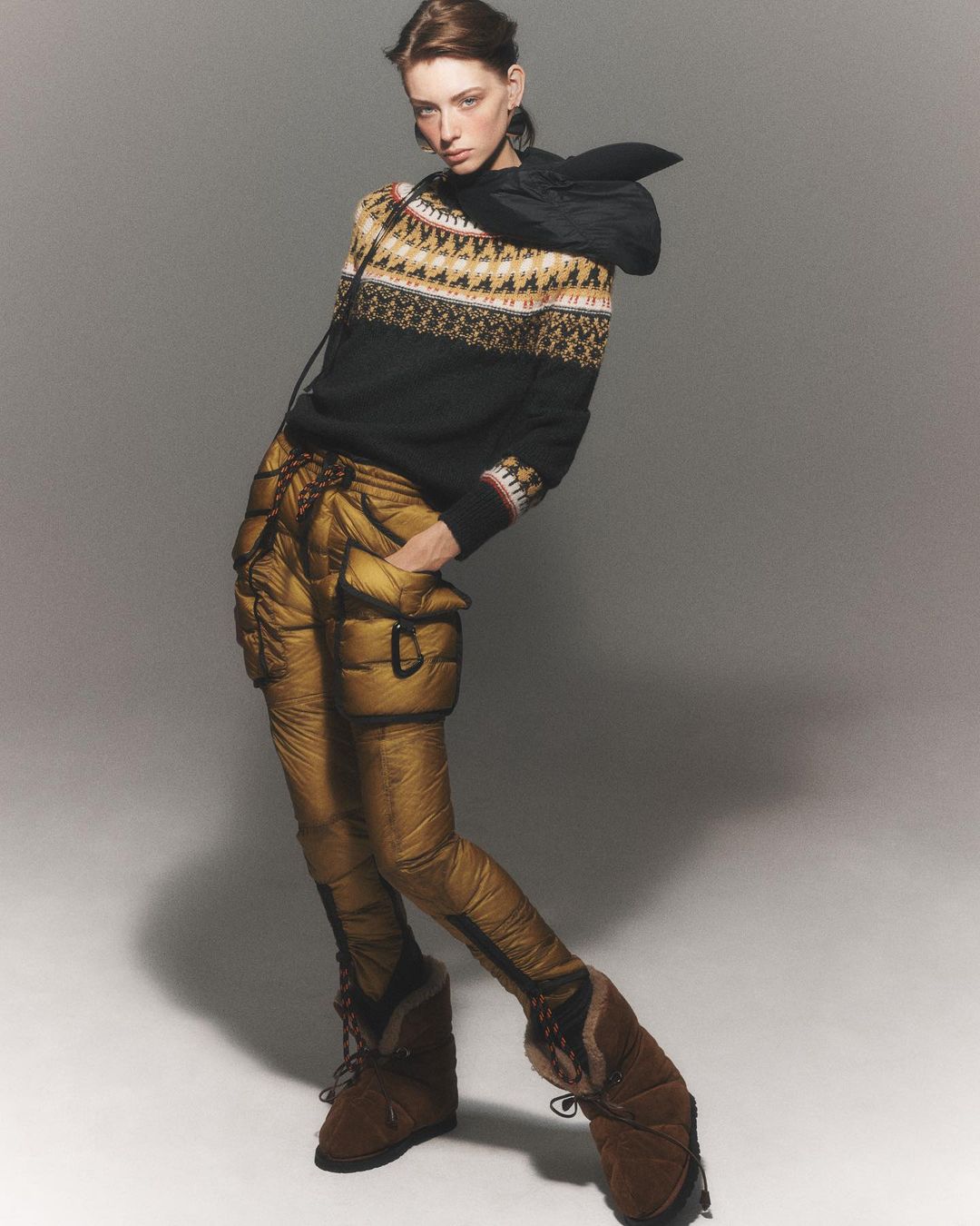 Full-length shot of a woman in gold puffer pants, a patterned sweater, and brown boots against a neutral studio backdrop.
