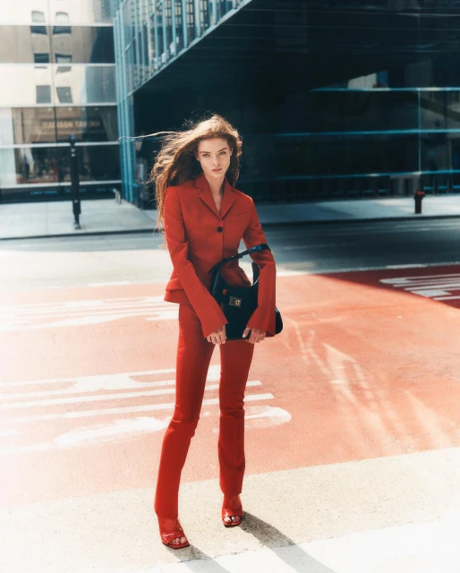 Fashionable woman in a red suit stands confidently on a crosswalk with modern buildings in the background, holding a black handbag.