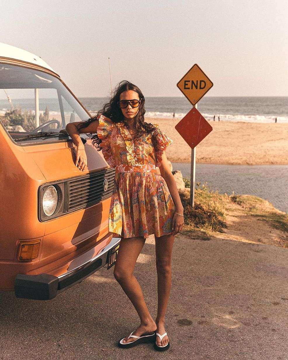 Adult female model poses next to an orange van near a beach with an 'End' sign, showcasing fashion in an outdoor setting.