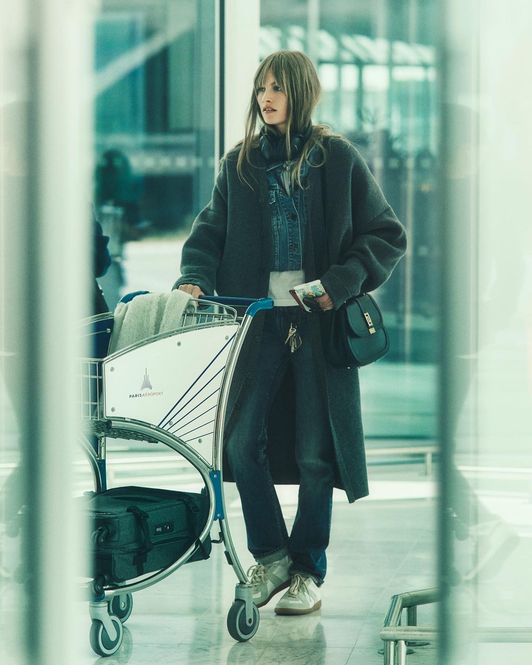 Adult woman in a long coat pulling an airport luggage cart in an airport terminal with large windows and bright, diffused light.
