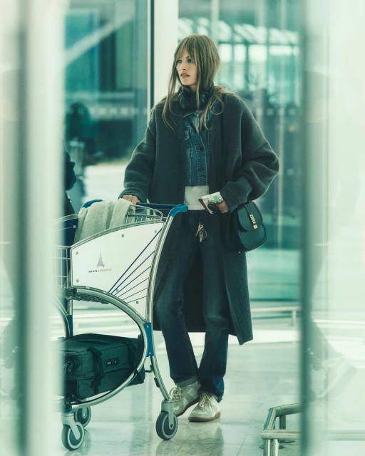 Adult woman in a long coat pulling an airport luggage cart in an airport terminal with large windows and bright, diffused light.