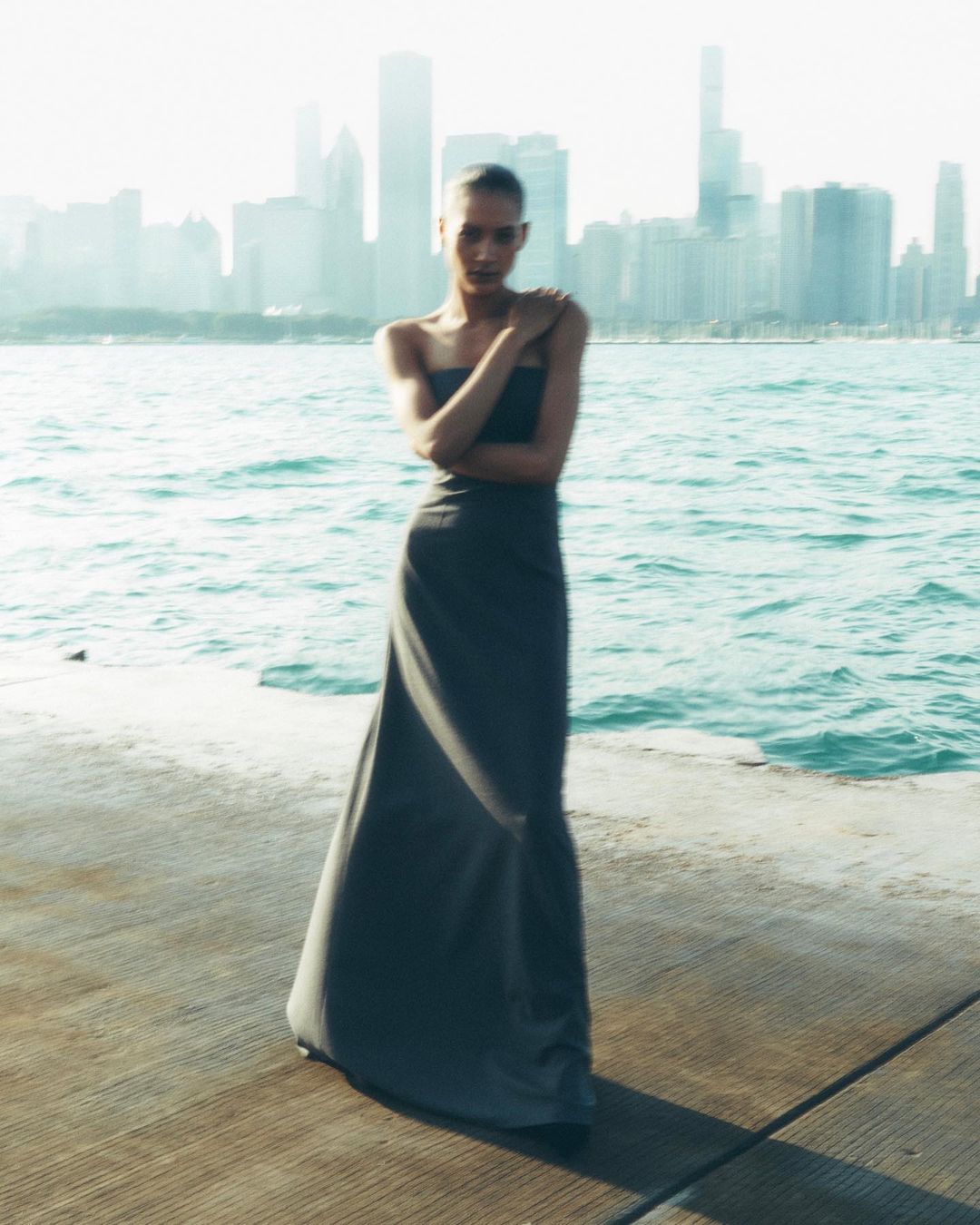 Woman in gray gown stands on concrete pier with Chicago skyline backdrop and turquoise water. Posing, fashion shoot, sunlight.