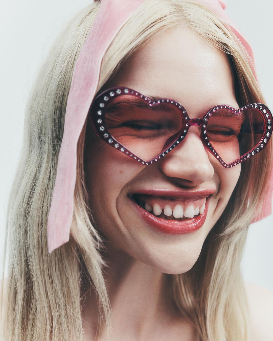 Close-up of a blonde woman with heart-shaped sunglasses adorned with gemstones and a matching pink headband, smiling against a white backdrop.