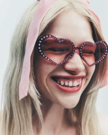 Close-up of a blonde woman with heart-shaped sunglasses adorned with gemstones and a matching pink headband, smiling against a white backdrop.