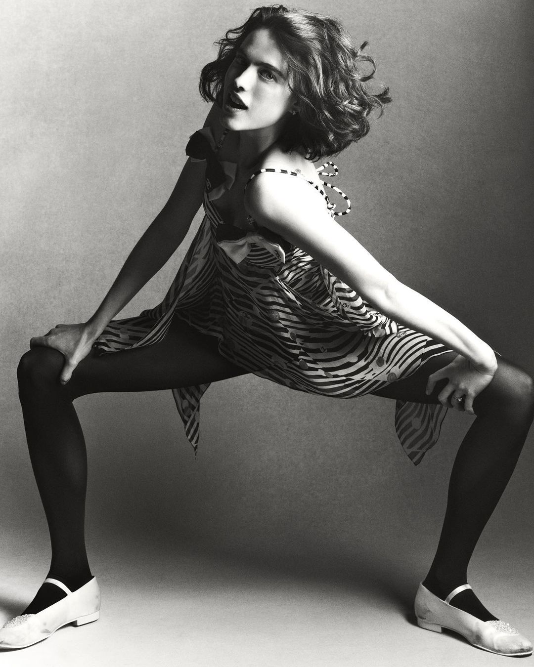 A young woman poses dynamically in a zebra-striped dress, black tights, and white shoes, set against a neutral grey backdrop in a studio setting.