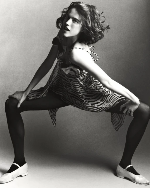A young woman poses dynamically in a zebra-striped dress, black tights, and white shoes, set against a neutral grey backdrop in a studio setting.