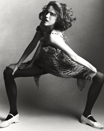 A young woman poses dynamically in a zebra-striped dress, black tights, and white shoes, set against a neutral grey backdrop in a studio setting.