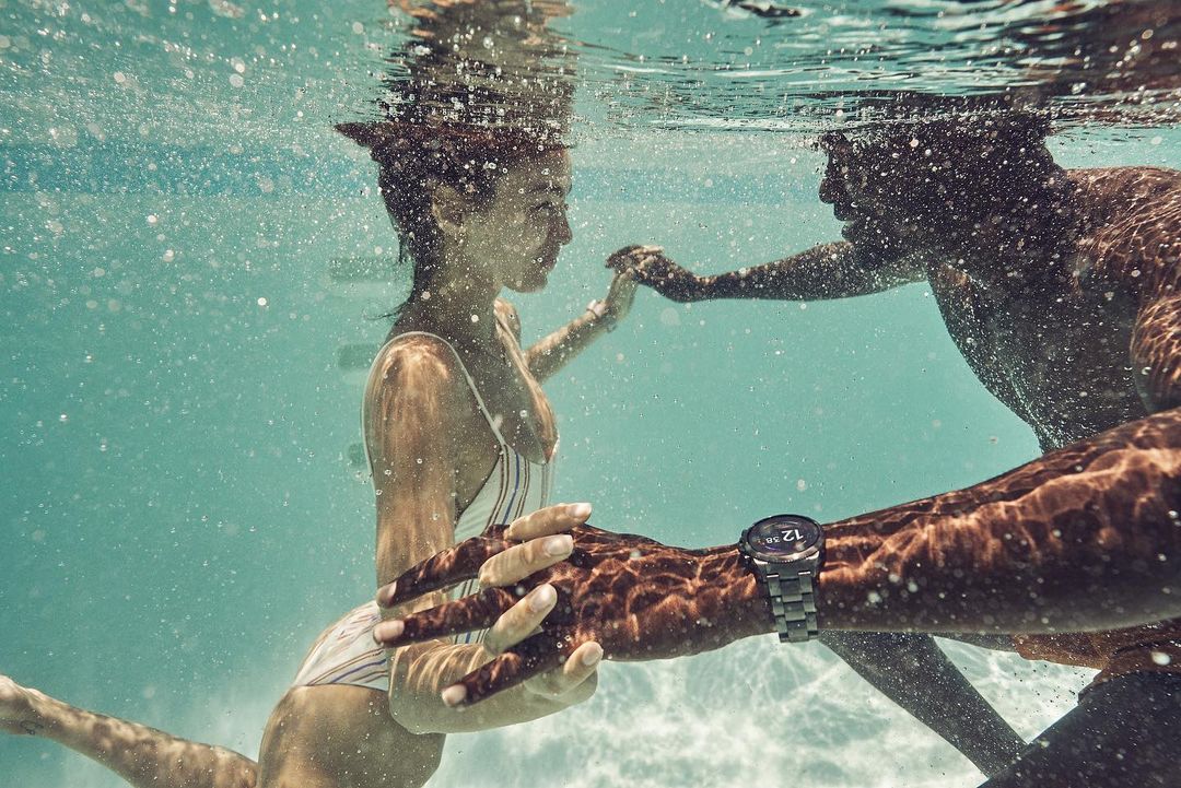 Underwater shot of a couple holding hands in a pool, with a man's arm and wrist visible in the foreground, wearing a smart watch.