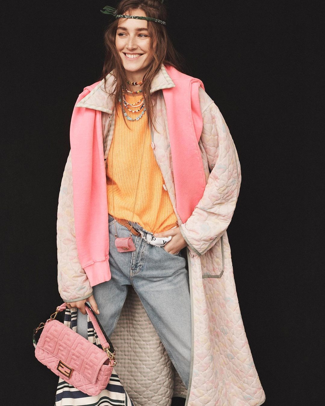 Smiling woman in a beige coat, orange sweater, and light-wash jeans stands against a black background, carrying a pink purse.