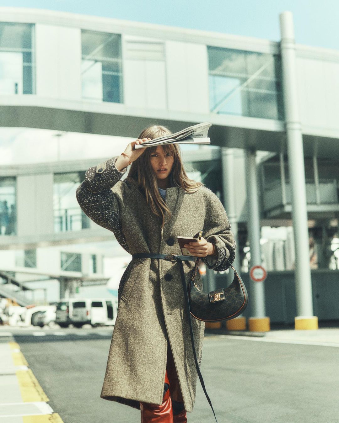 A woman in a belted coat poses outdoors with a newspaper on her head, holding a small bag in daylight, with airport building background.