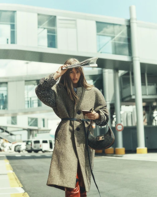 A woman in a belted coat poses outdoors with a newspaper on her head, holding a small bag in daylight, with airport building background.