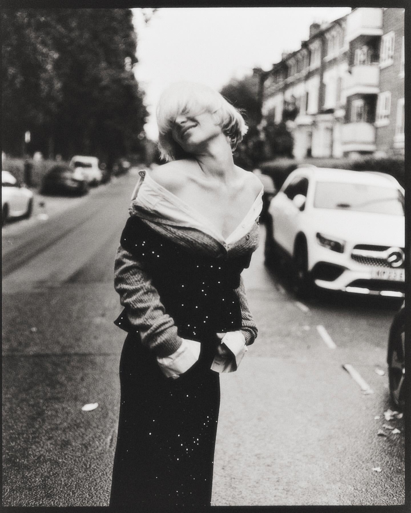 Monochrome portrait of a woman in an off-the-shoulder top and skirt standing on a street with trees and parked cars.