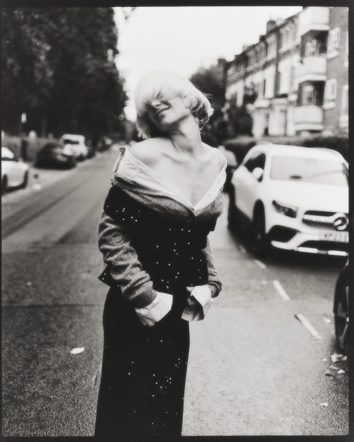 Monochrome portrait of a woman in an off-the-shoulder top and skirt standing on a street with trees and parked cars.