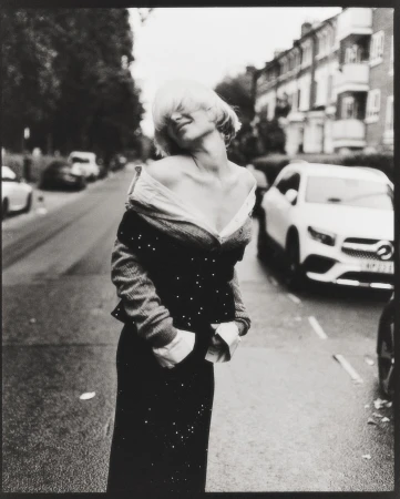 Monochrome portrait of a woman in an off-the-shoulder top and skirt standing on a street with trees and parked cars.
