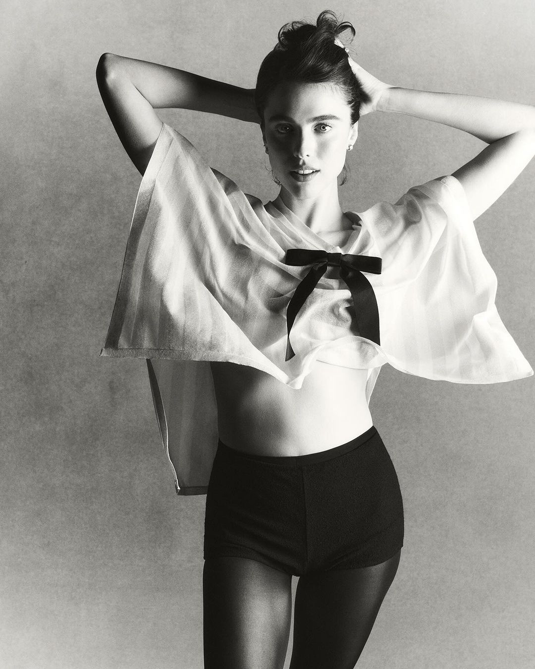 Black-and-white portrait of a fair-skinned woman wearing a sheer white top with a black bow, shorts, and stockings, with hands in her hair.