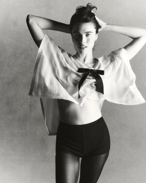 Black-and-white portrait of a fair-skinned woman wearing a sheer white top with a black bow, shorts, and stockings, with hands in her hair.